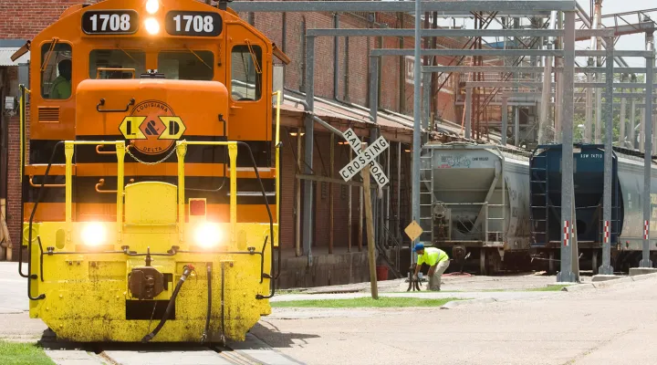 Train pulling covered hopper cars on tracks that are street level with a brick building in the background next to a railroad crossing sign