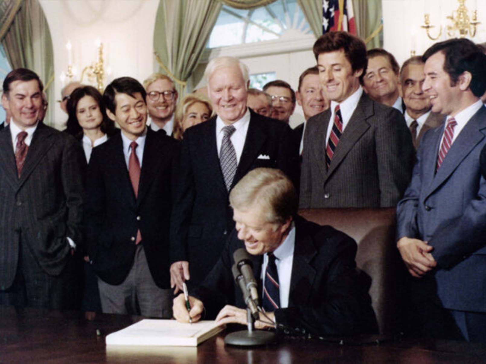 President Jimmy Carter sitting down at his desk, signing a document with a group of people standing behind him
