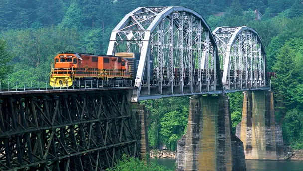Orange and yellow train with cargo riding through a bridge over water with trees in the background.