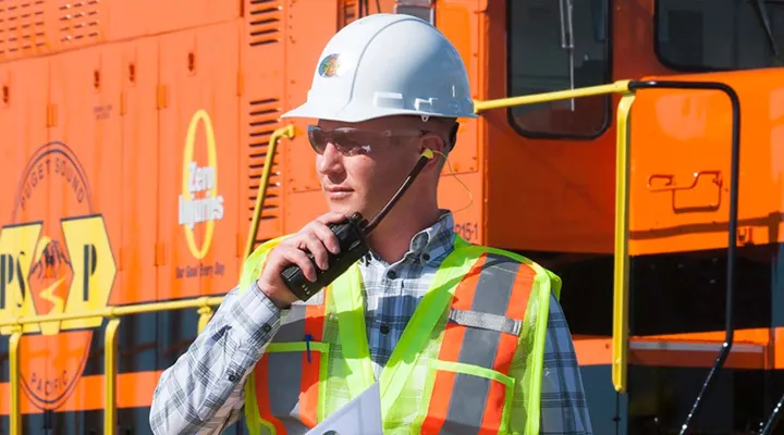 G&W employee wearing a safety vest, glasses, and helmet with a train in the background