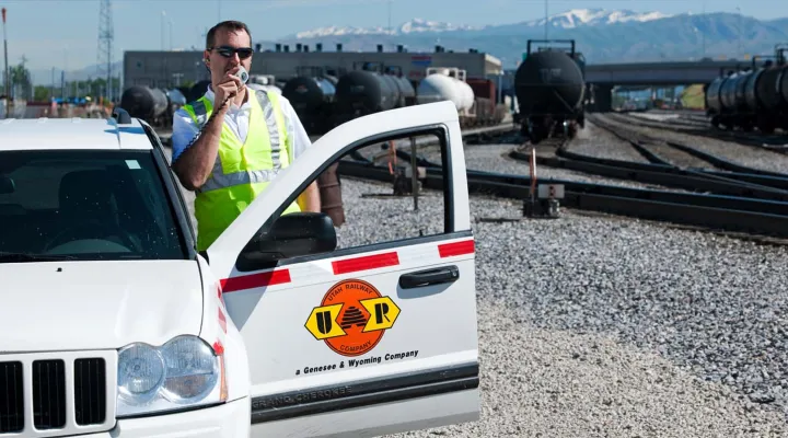 G&W employee in a high-visibility vest and sunglasses standing outside of a Genesee & Wyoming car who is talking into a walkie-talkie
