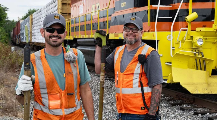 Two G&W employees wearing high-visibility vests and smiling with a train in the background